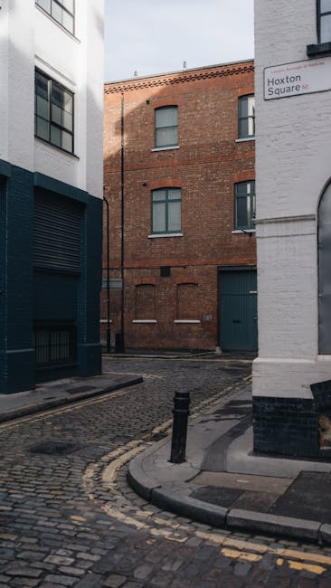 The image depicts a narrow urban street corner in Hoxton, with a small section of cobbled pavement bordered by a low curb and two short black bollards. On the left, there is a modern white building wi