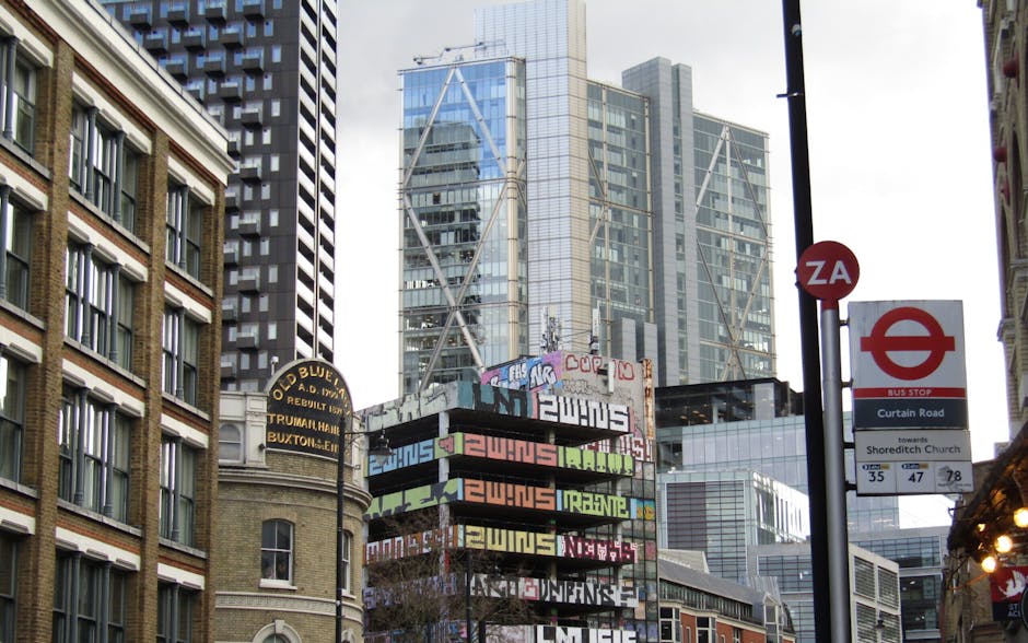The image depicts a narrow urban street corner in Hoxton, with a small section of cobbled pavement bordered by a low curb and two short black bollards. On the left, there is a modern white building wi