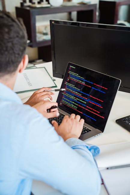 A person sitting at a desk is engaged in coding or debugging on a laptop, which displays lines of multicolored programming code on a dark background. The individual, seen from behind and wearing a lig